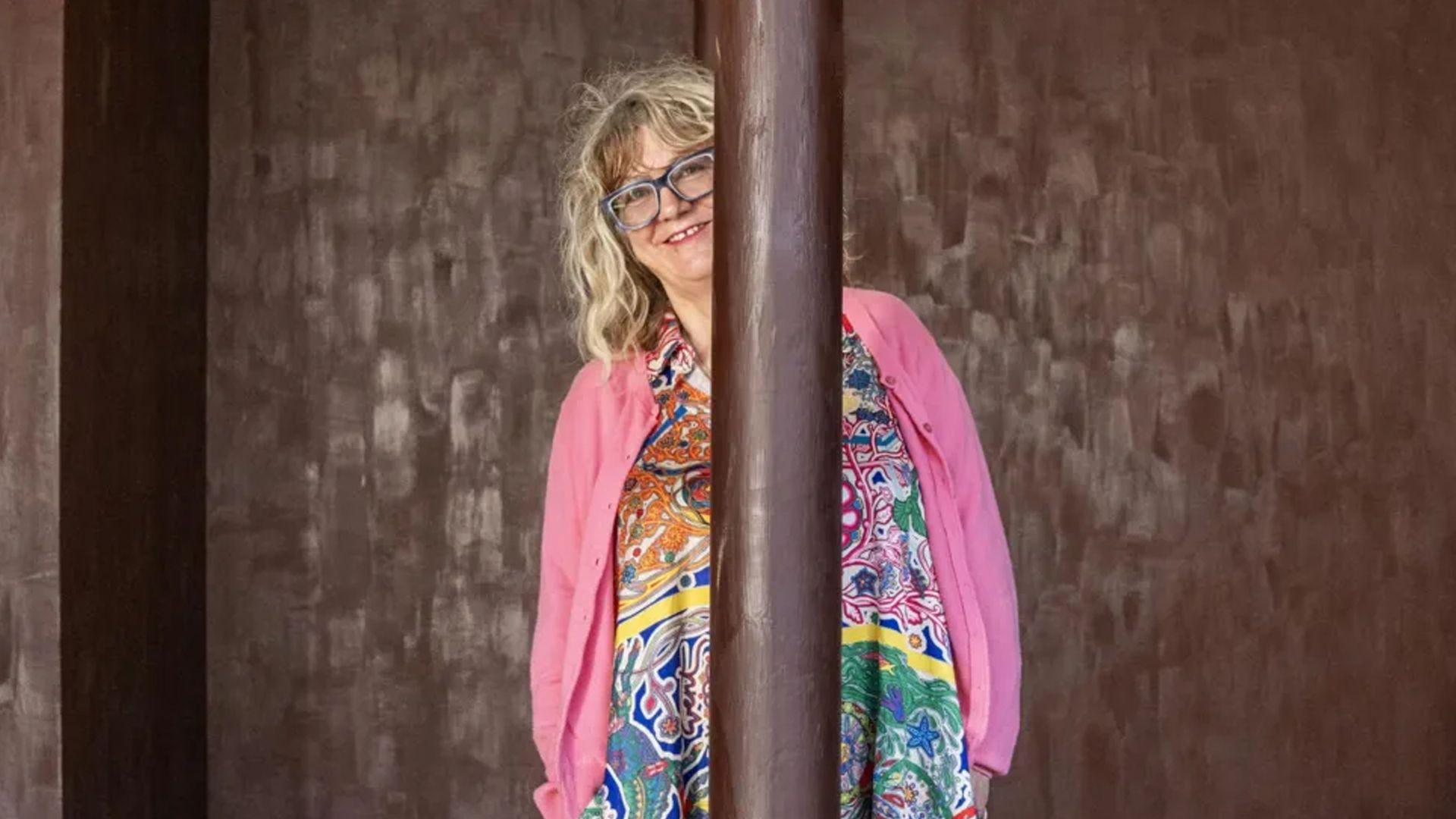 A woman wearing a bright coloured, patterned dress and pink cardigan behind a pillar painted with melted chocolate