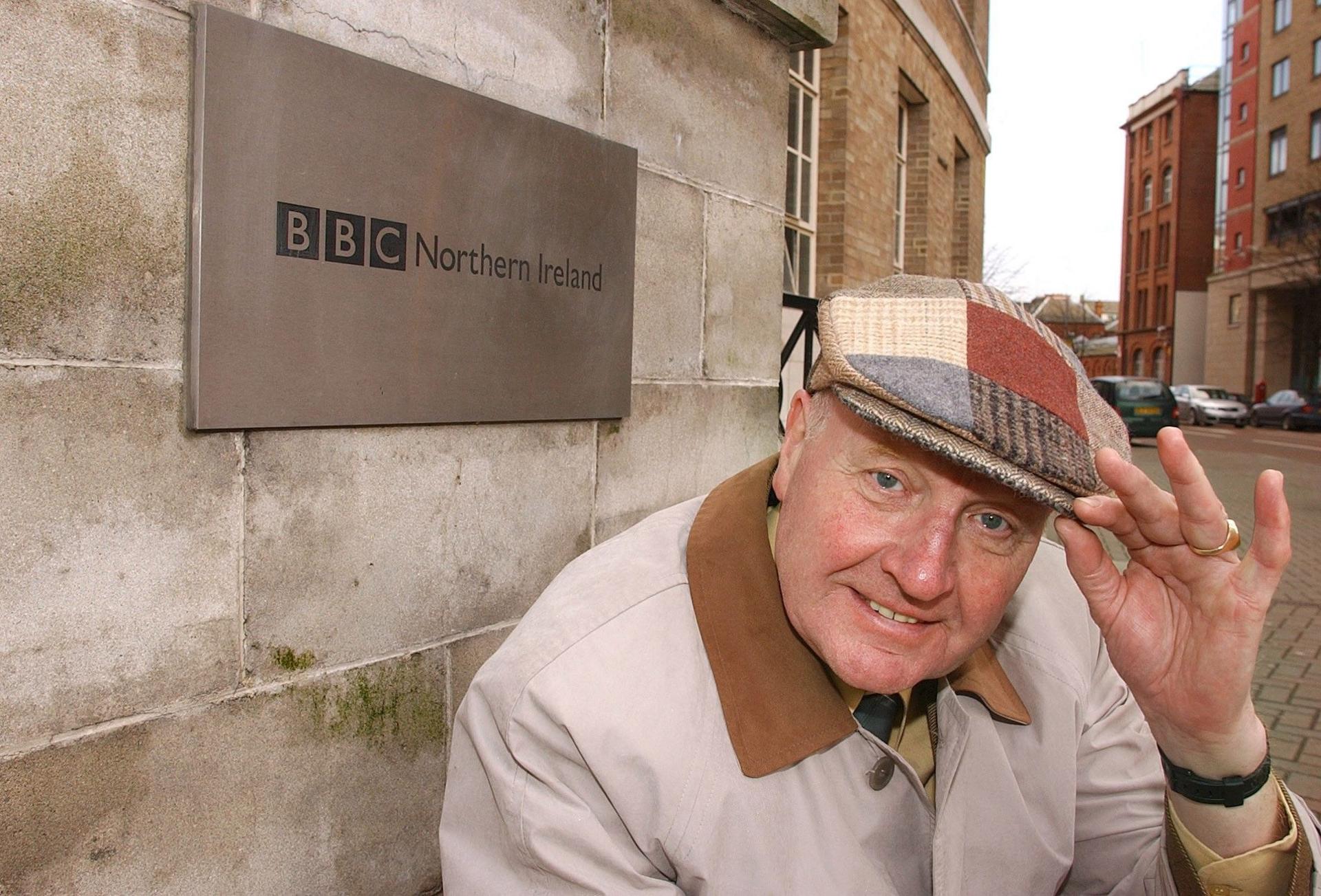 John Bennett in a beige coat, hand raised to a patterned flat cap, stood beside BBC Northern Ireland building with signage