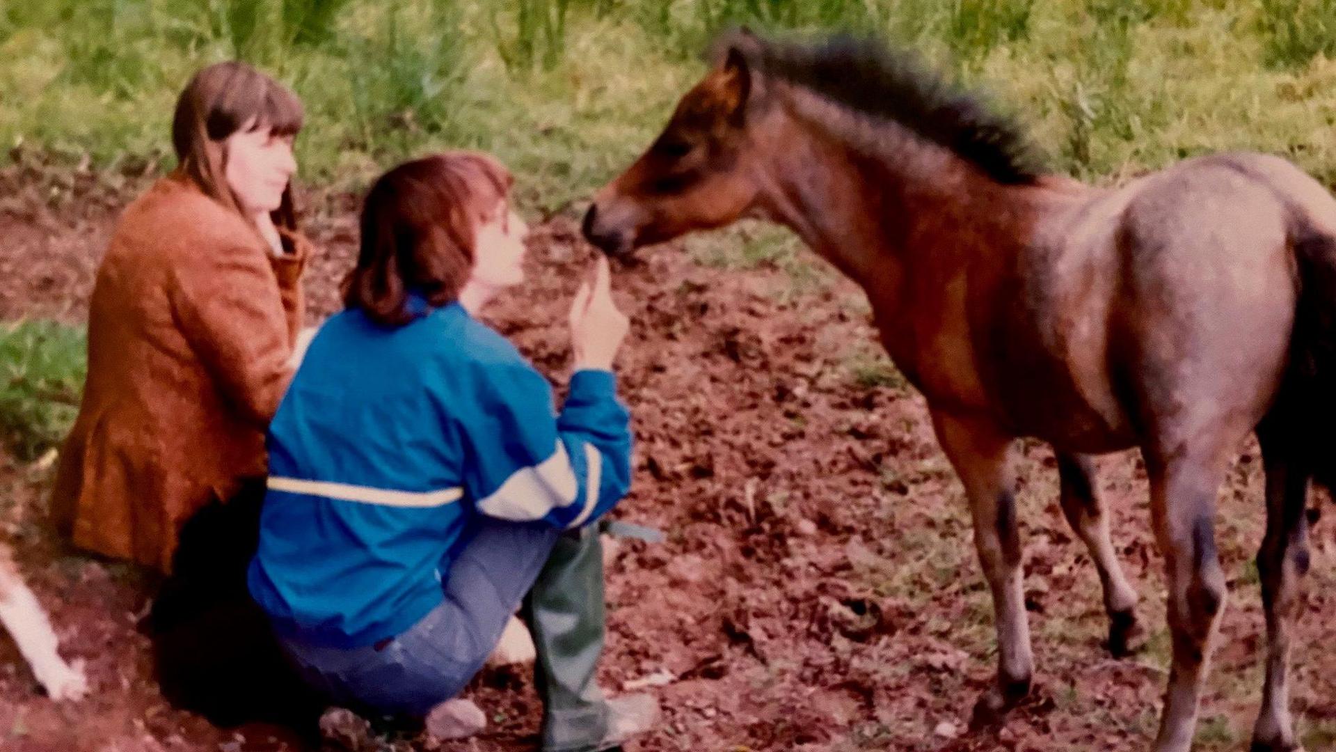 Ginny and Stella sitting on the ground next to a small brown foal. Both have a fringe and dark hair, and are turned away from the camera towards the foal. Ginny is wearing a brown jacket while Stella is wearing a bright blue jacket. 