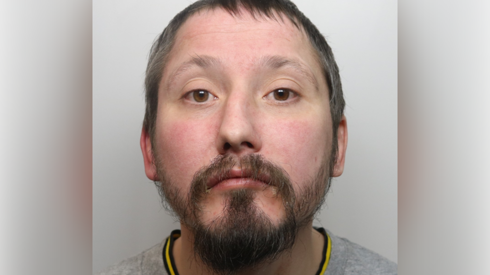 A police mugshot of a man with short black hair, a black beard and a grey top.