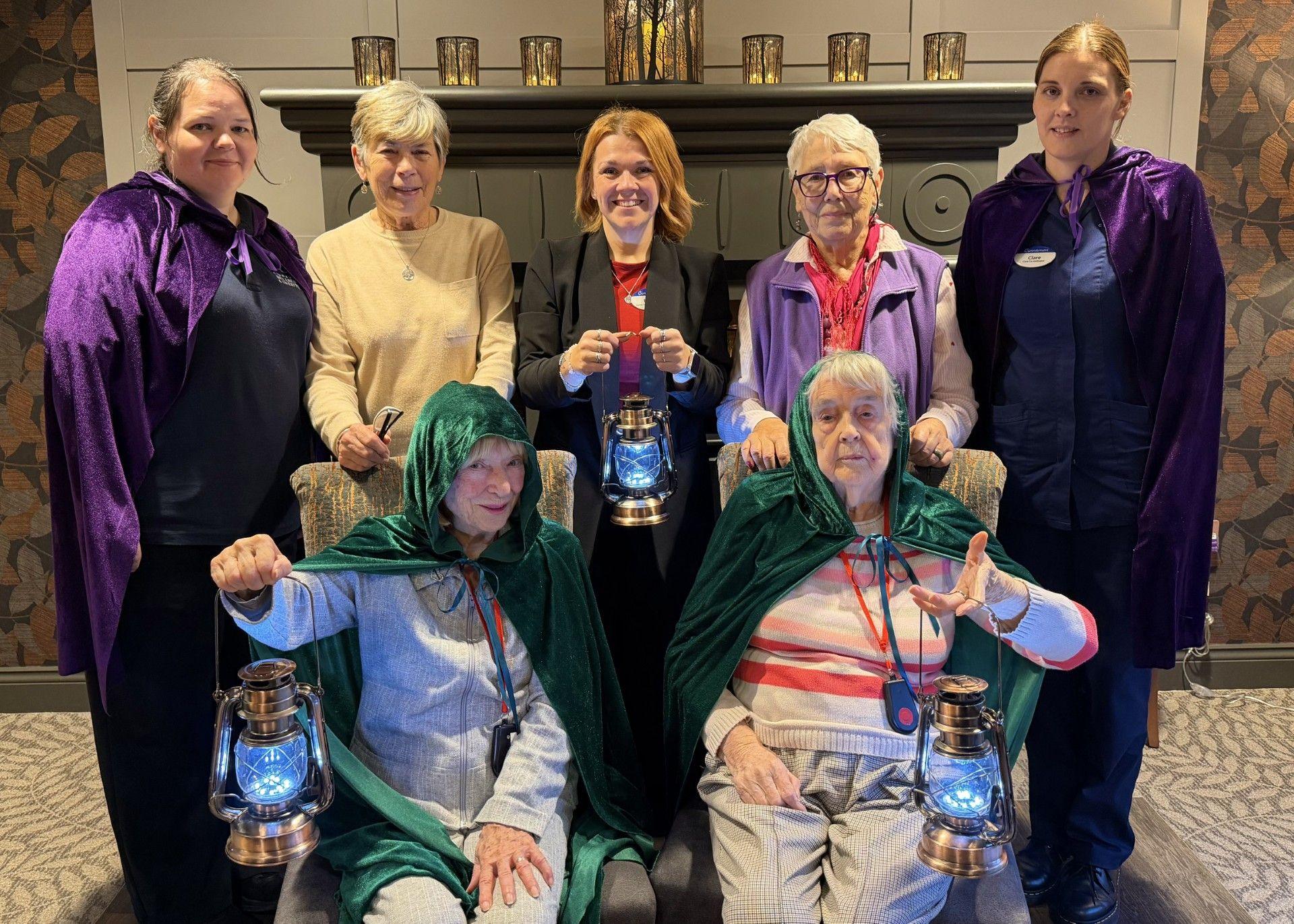 Seven members of staff and residents of Parkfield Grange Care Home can be seen wearing blue and green cloaks. Three of them are holding up lamps in the style of the Traitors television programme. 