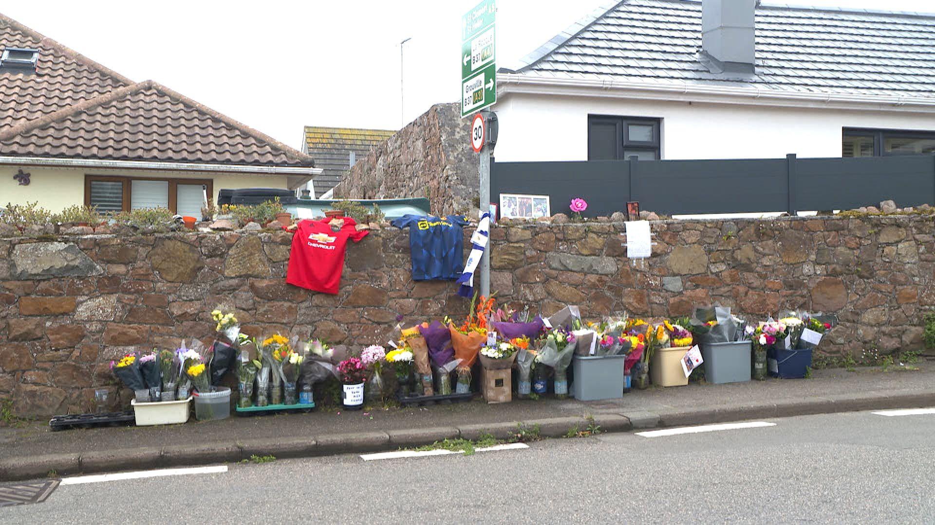 A photo of La Rue de Fauvic in Grouville, with flowers lined up in tribute along the pavement. There are two football shirts on a stone wall, with houses behind the wall.