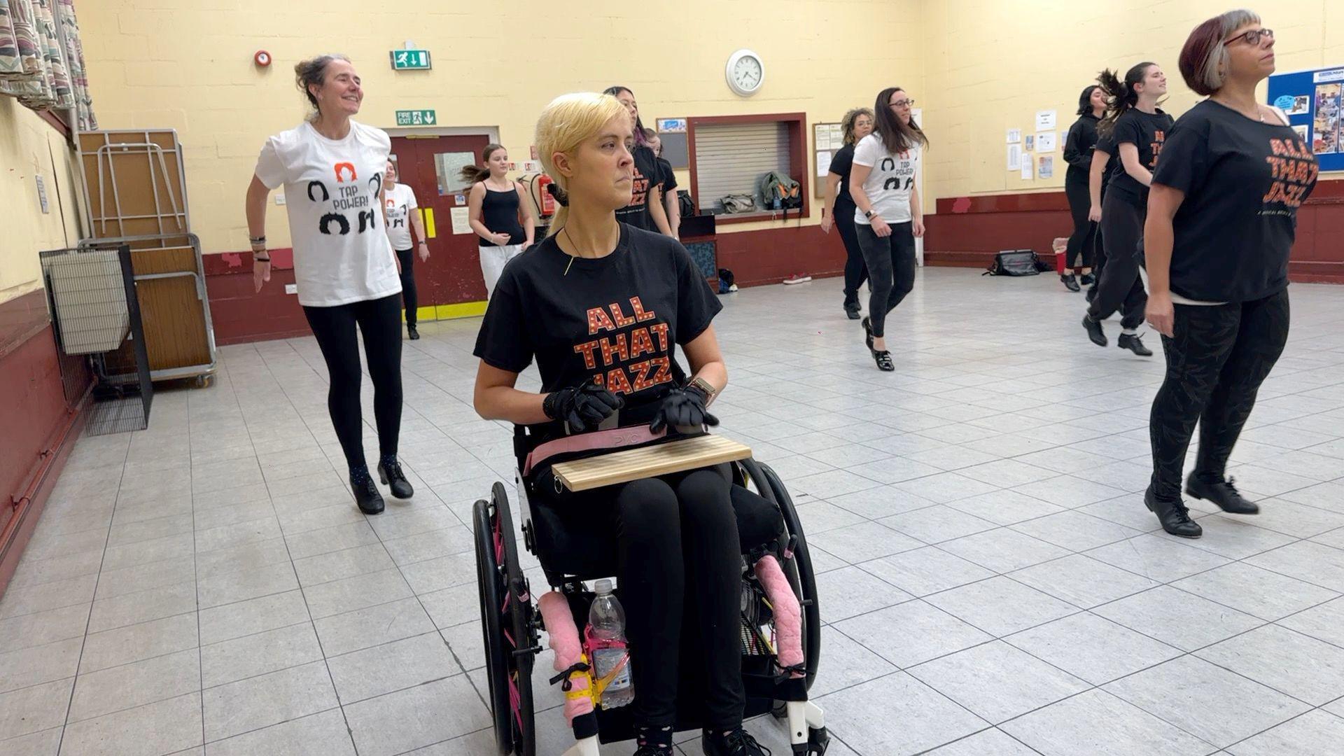 Ms Booth is on the front row of a group of tap dancers who are practising in a dance studio. 