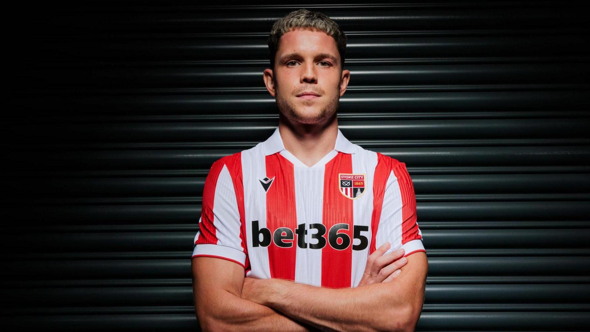 George Hirst poses with his arms crossed in Stoke's red-and-white striped home shirt