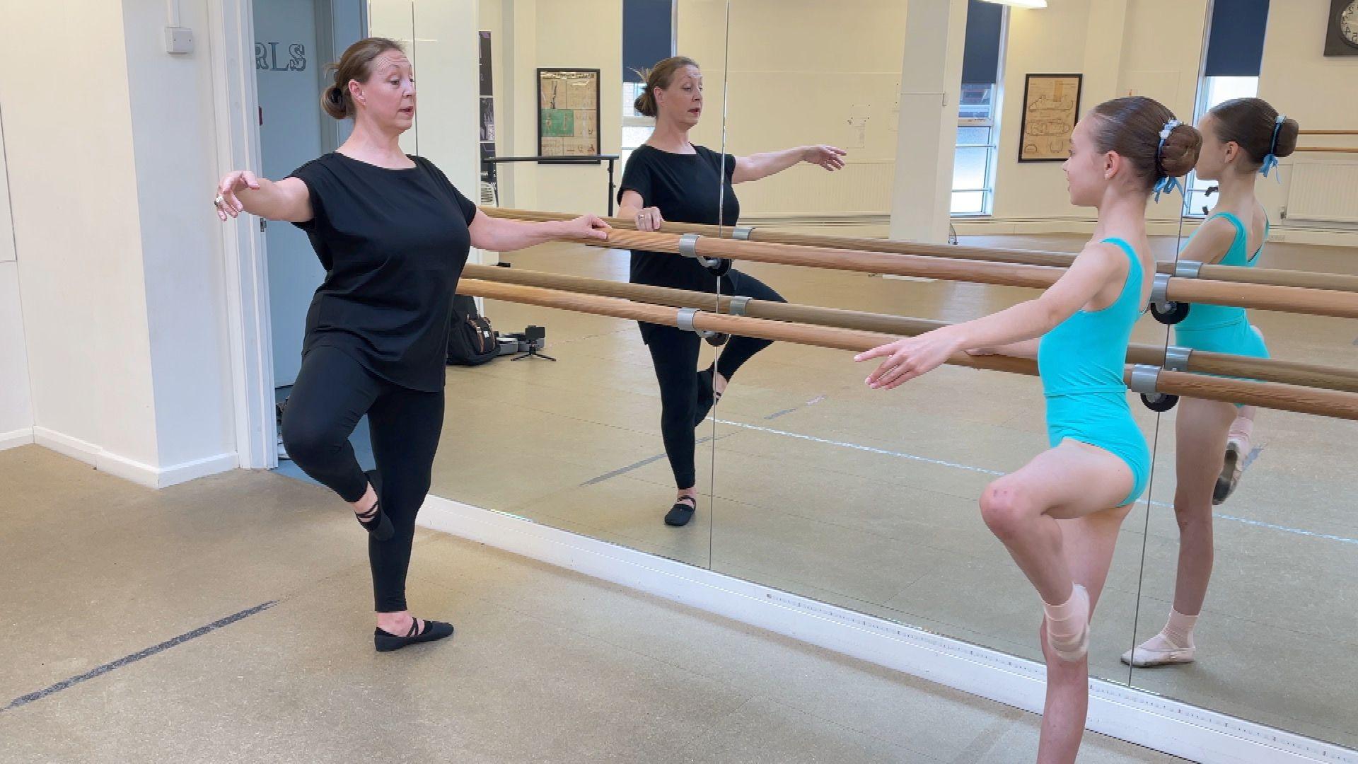 Trina Lee in black clothing doing a passé ballet, while holding on to the wooden bar attached to the mirror. Opposite is Jessica, in her blue leotard, mimicking the move.
