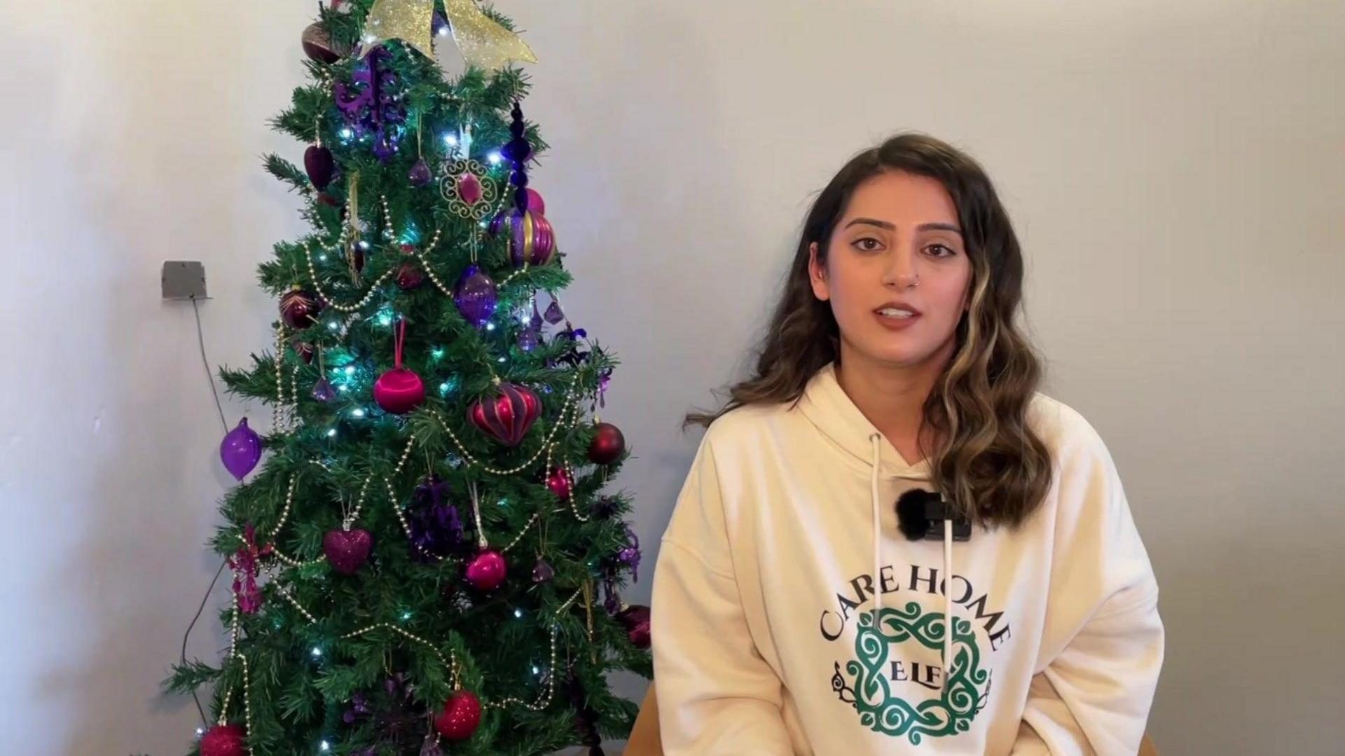 Farah is standing next to a decorated Christmas tree. She is wearing a white hoodie which says 'care home elf'.