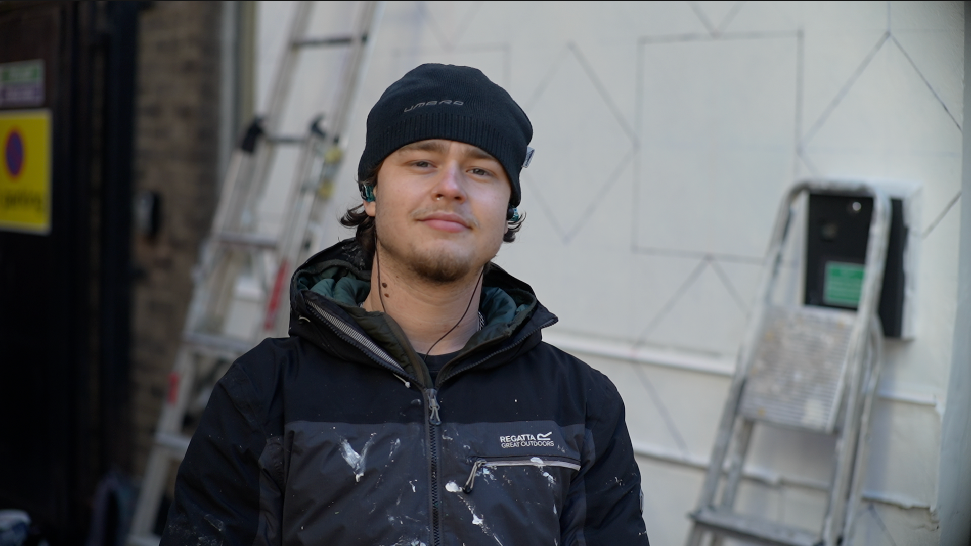 Leo Vanmegan smiles at the camera as he stands in front of a white building with some shapes drawn on to it and a step ladder that leans against it. He wears a black beanie hair, black coat and has wired headphones in each ear. He has dark hair that pokes out of his hat and he has a beard.