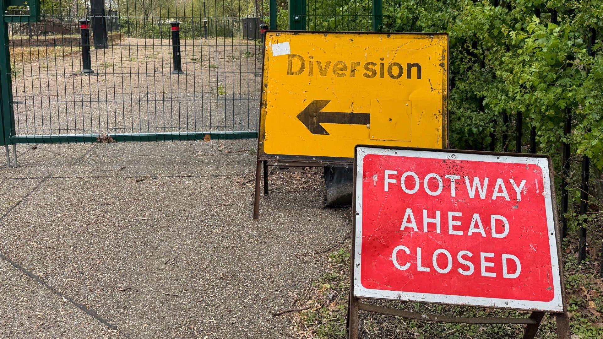 A red road sign with the words "FOOTWAY AHEAD CLOSED" in white lettering in front of a yellow road sign with the word "Diversion" above a black arrow. Both signs are on a pavement in front of a bridge which is fenced off to the public.