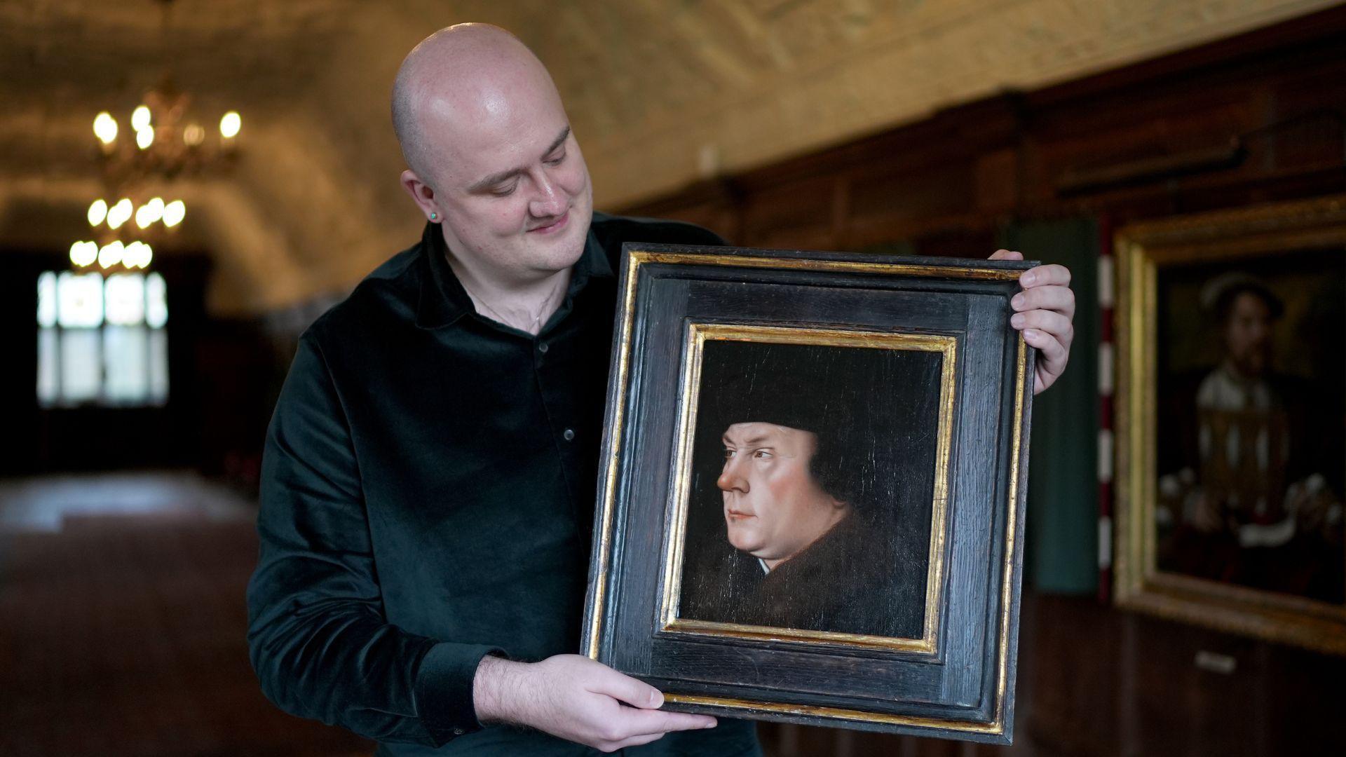 A bald white man wearing a black shirt. He is holding a framed portrait of Thomas Cromwell. The man is stood in a long room with brown wooden walls and a cream ceiling. He is looking down and to the right at the picture he is holding