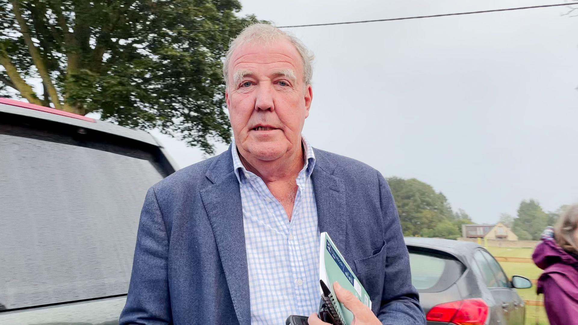 Jeremy Clarkson wears a navy blazer and white and blue checked shirt as he stands behind his car in a rainy countryside car park, holding a notebook and his phone ahead of a council meeting.