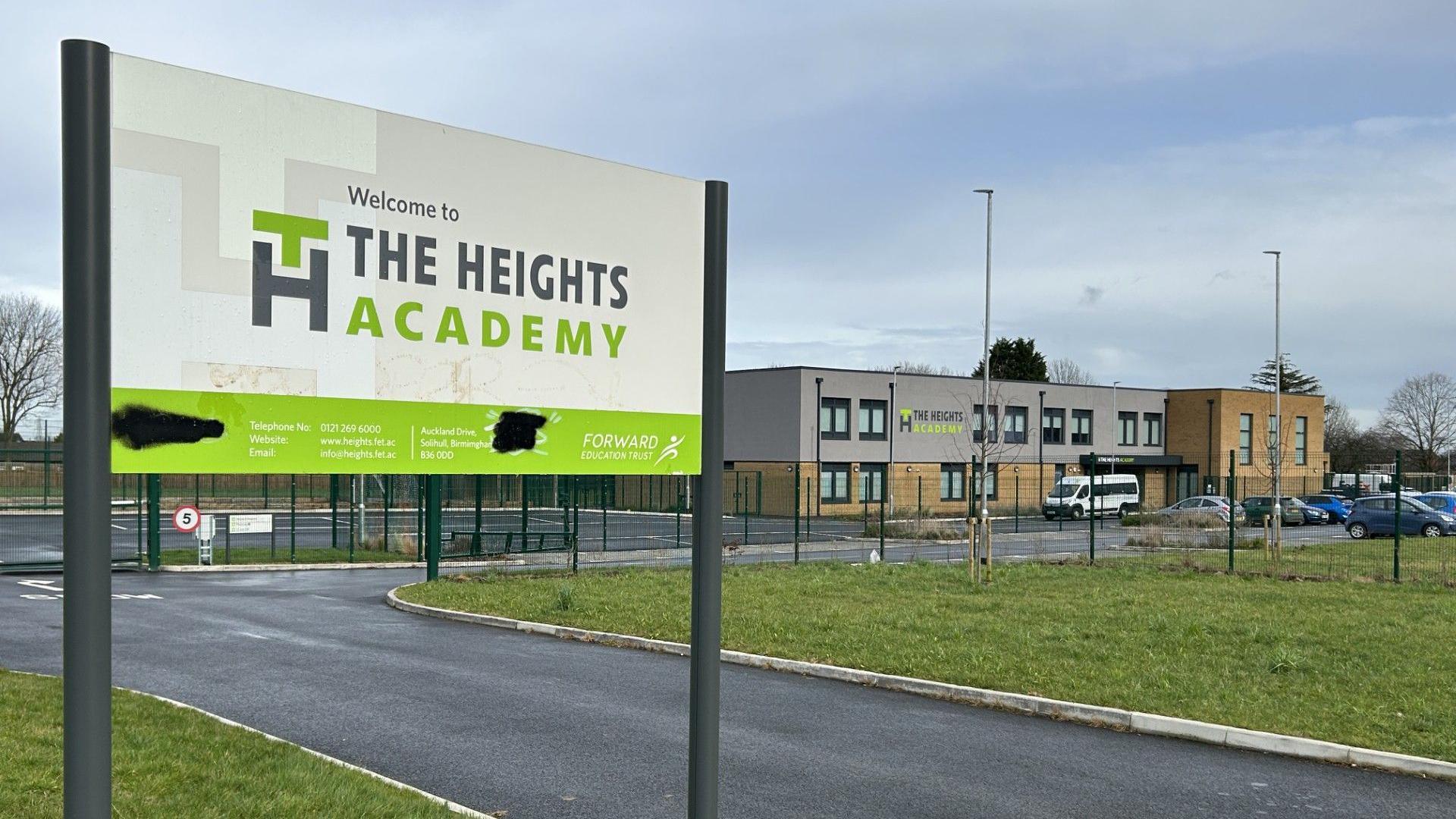 The Heights Academy in Smith's Wood has a white and green sign at the entrance to the school. In the background is the two-storey building which hasa flat roof and a service road providing access. A car park at the front of the building can also be seen. 