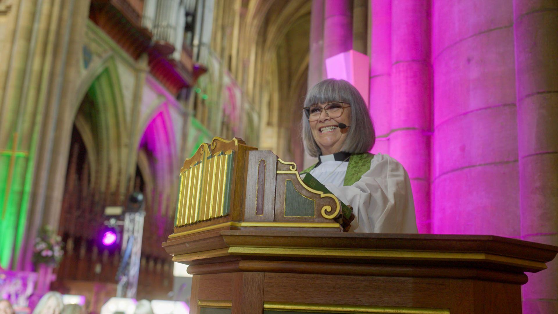 Dawn French is standing behind a brown lectern. She is wearing a cassock and is looking out at the congregation and smiling. Her hair is grey.