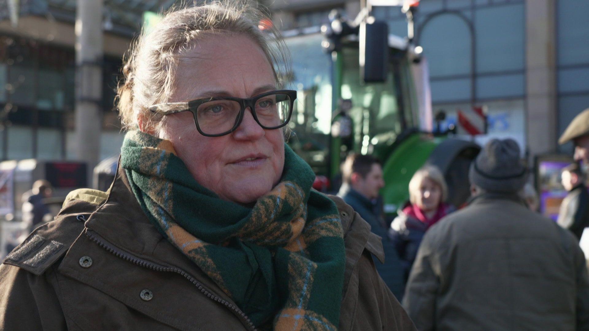 Rachel Hallos stands in front of a tractor in Chester's Town Hall Square. She is wearing a brown coat and green and yellow scarf and is wearing glasses.