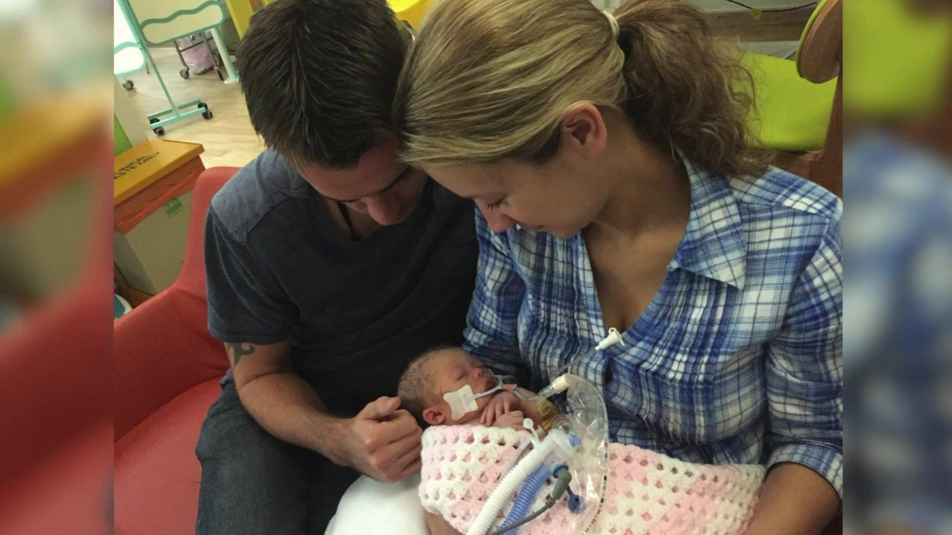 A man and a woman holding a baby wrapped in a pink and white blanket. They are looking down and smiling. 