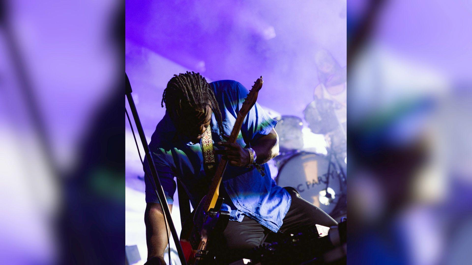 Lead singer nand guitarist for Bloc Party, Kele Okereke, is performing on stage. He is leaning over his electric guitar as he plays, the background is purple and he wears a purple short sleeved shift with black trousers.