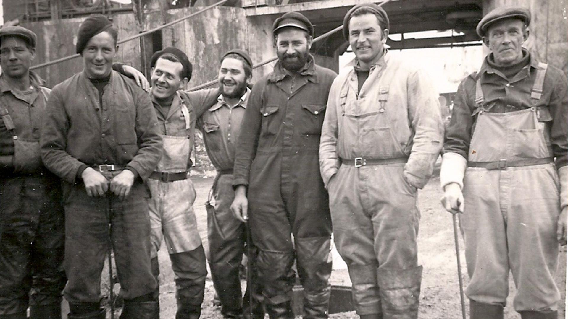 A group of seven whalers posing for the camera in a black and white picture. All are looking at the camera except the man in waders third from the left, who is staring at a man in overalls in a hat. The man on the near right is wearing waders and is leaning on a stick.