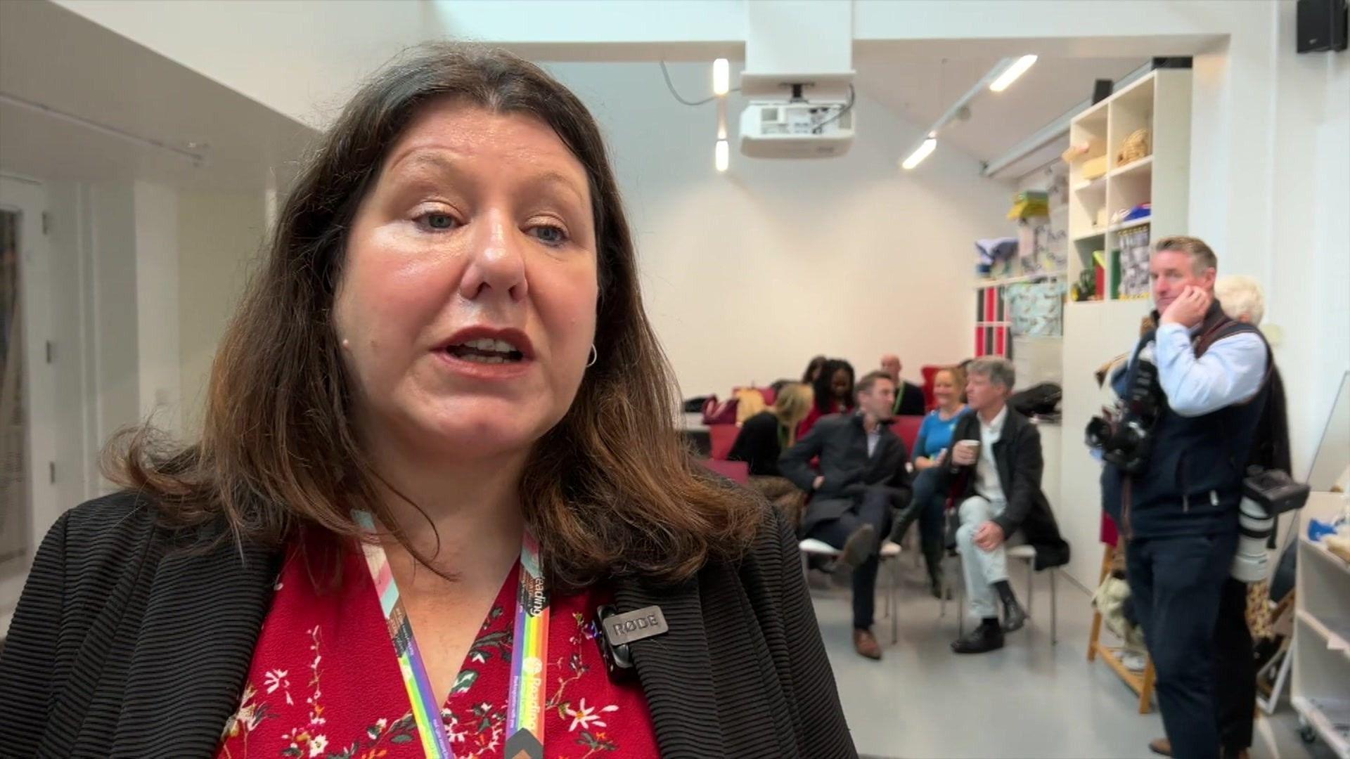 A woman with shoulder length brown hair wearing a red blouse, a black blazer and a rainbow lanyard. The inside of a hall with a small crowd of people sitting on chairs can be seen behind her.