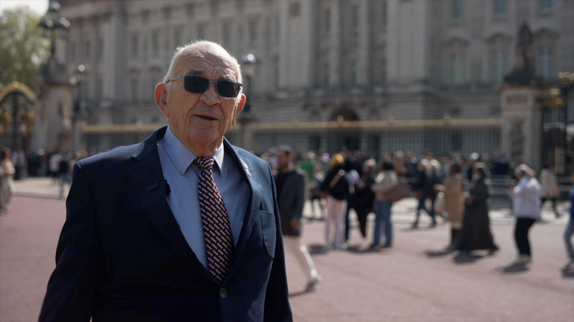 Bob is stood outside Buckingham Palace, wearing a navy suit, blue shirt and red tie. He has short white hair and is wearing dark sunglasses.