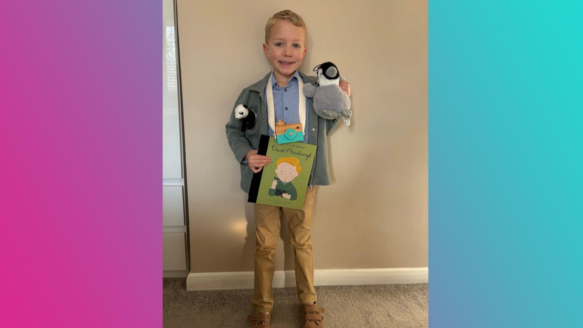 A boy smiling as he wears a blue shirt, a grey jacket, beige trousers and brown shoes. He's wearing a camera and is holding a toy bird, with a toy panda tucked into his jacket pocket.