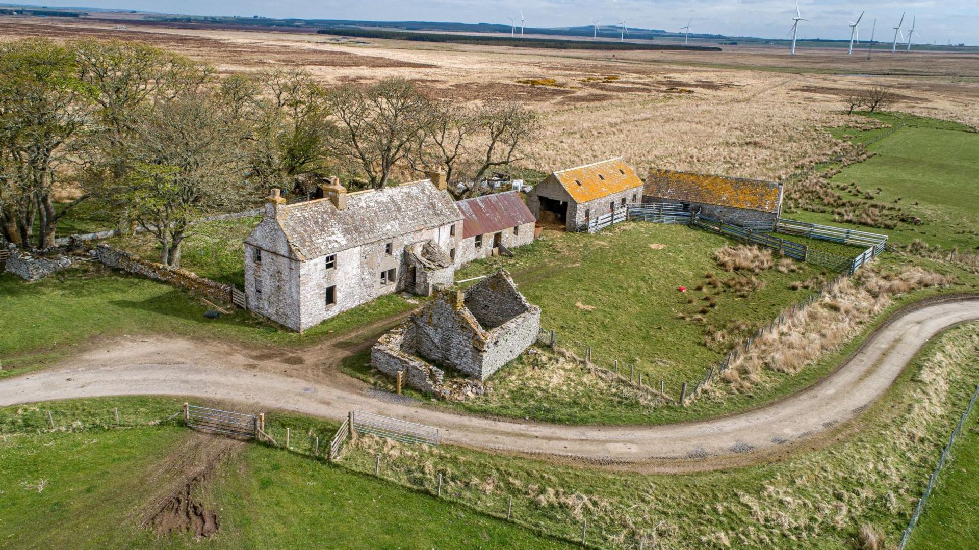 An aerial image of the farm house and outbuildings. There is a road that snakes by the property. Behind the farm house are trees. 