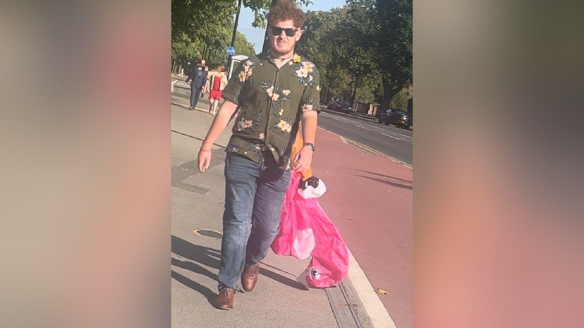 An image of a man with curly, ginger hair and a beard. He is wearing a khaki shirt with a yellow floral print, blue jeans and brown shoes. An inflatable pink flamingo costume appears to be hanging off his back.