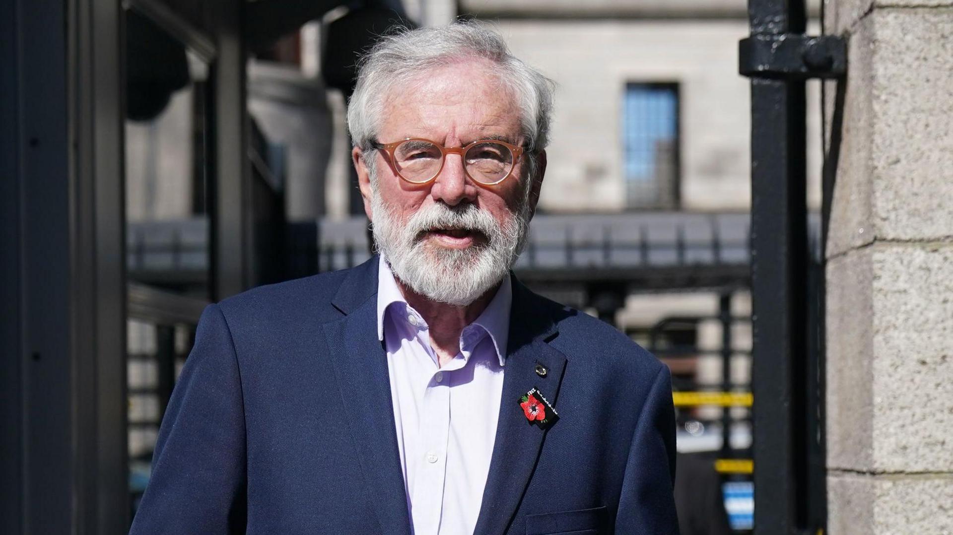 Gerry Adams, with grey hair and a grey beard, wearing a navy suit, a light pink shirt with the top button undone. He is wearing round orange glasses. He is walking through a black metal gate outside the High Court.