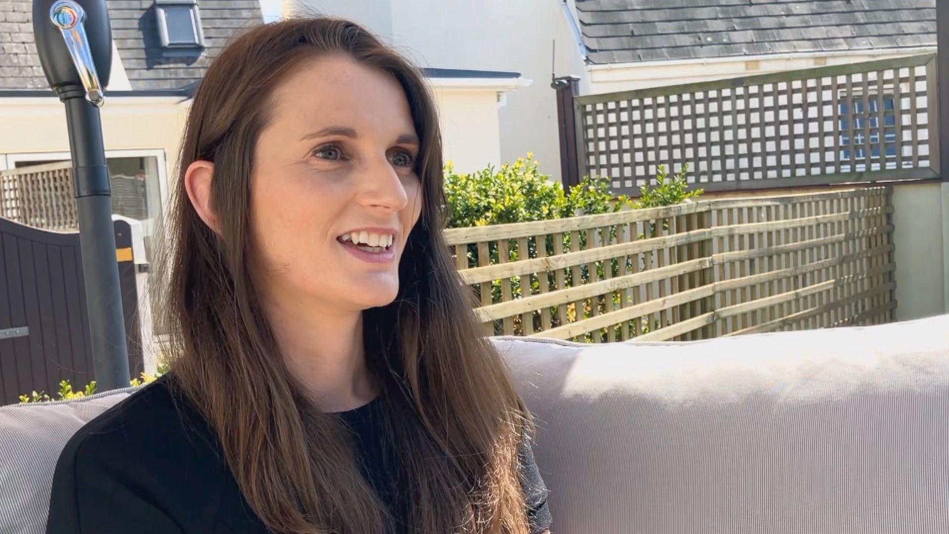 Laura has long brown hair and sits on an outdoor couch. Behind her is a wooden fence, green bushes, and parts of neighboring houses with grey roofs. A black lamp post stands on the left side of the image.
