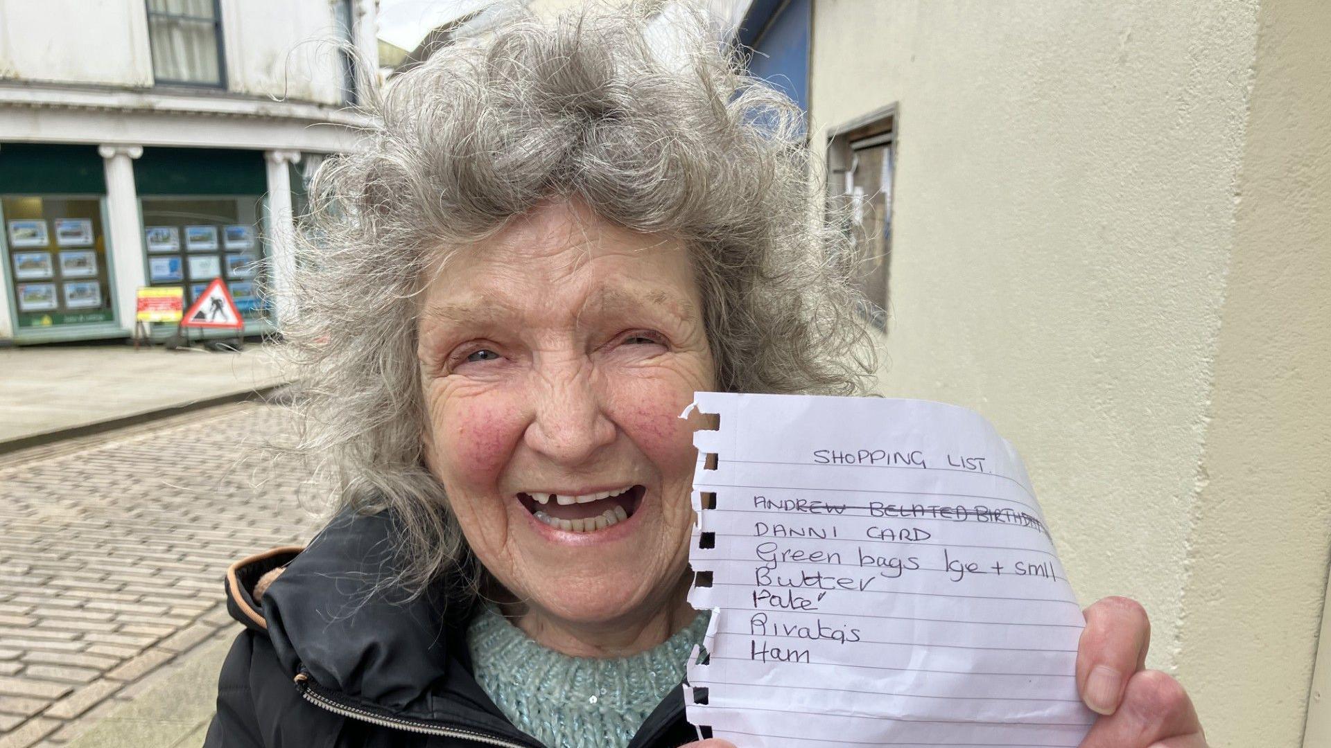 Irene Widger - an older lady with grey curly hair in standing in a street and laughing into the camera. She is holding a shopping list close to her face.
