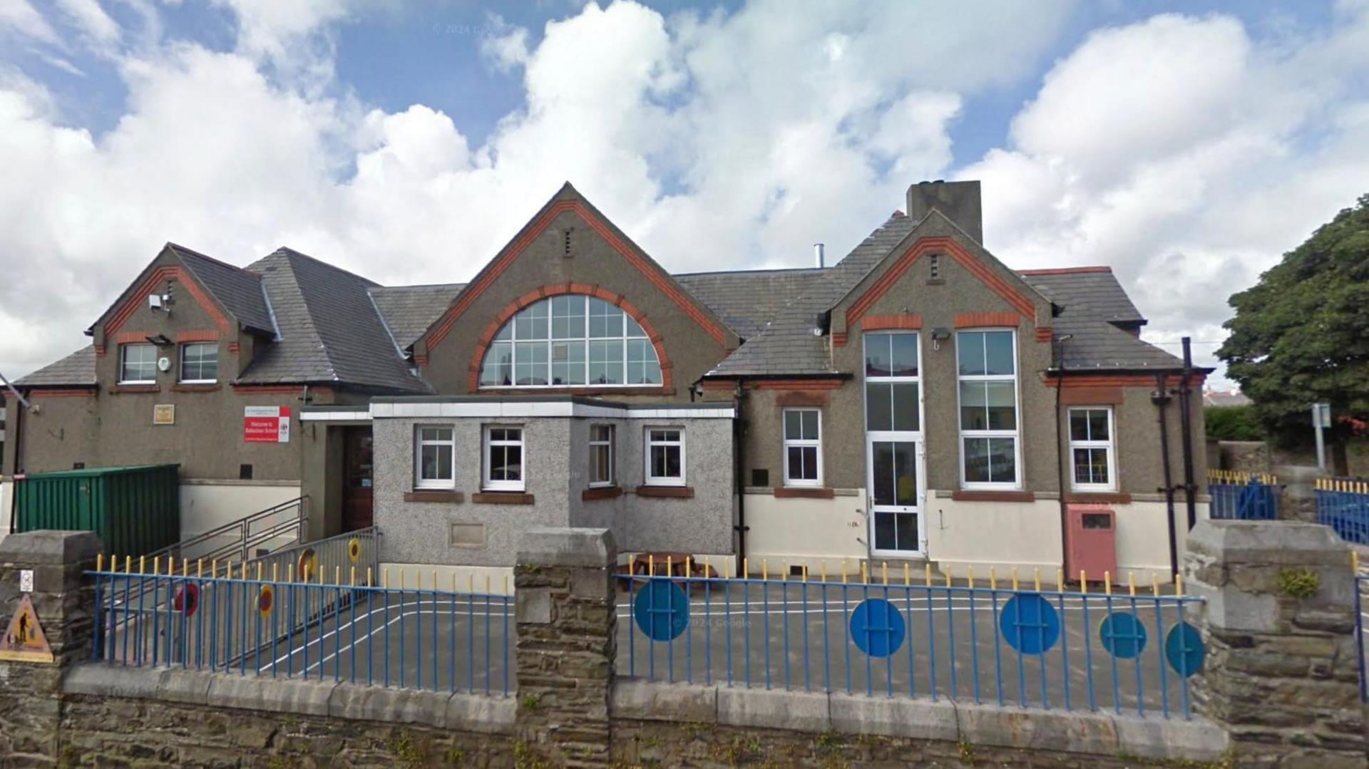 The exterior of Ballacloan School. It is a grey-brown cement building with a small playground outside, and blue railings to delineate the small school.