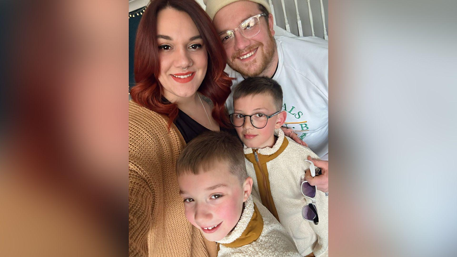 A family selfie-style photo, with Natasha and Joe at the back and their boys in front. Natasha has dyed brown hair, a nose ring and has red lipstick on. She is wearing a beige cardigan and black top. Joe has glasses and a beard, and is wearing a beige winter hat and white jumper. The boys have blond hair and Oscar is wearing glasses. They have matching beige fleeces.