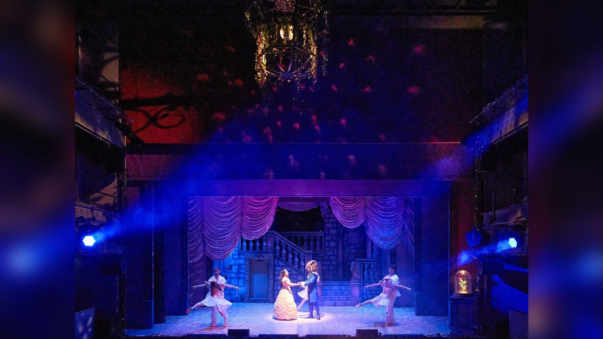 A theatre stage with actors and actresses performing a show. The stage is lit up and hanging from the ceiling is a chandelier. 