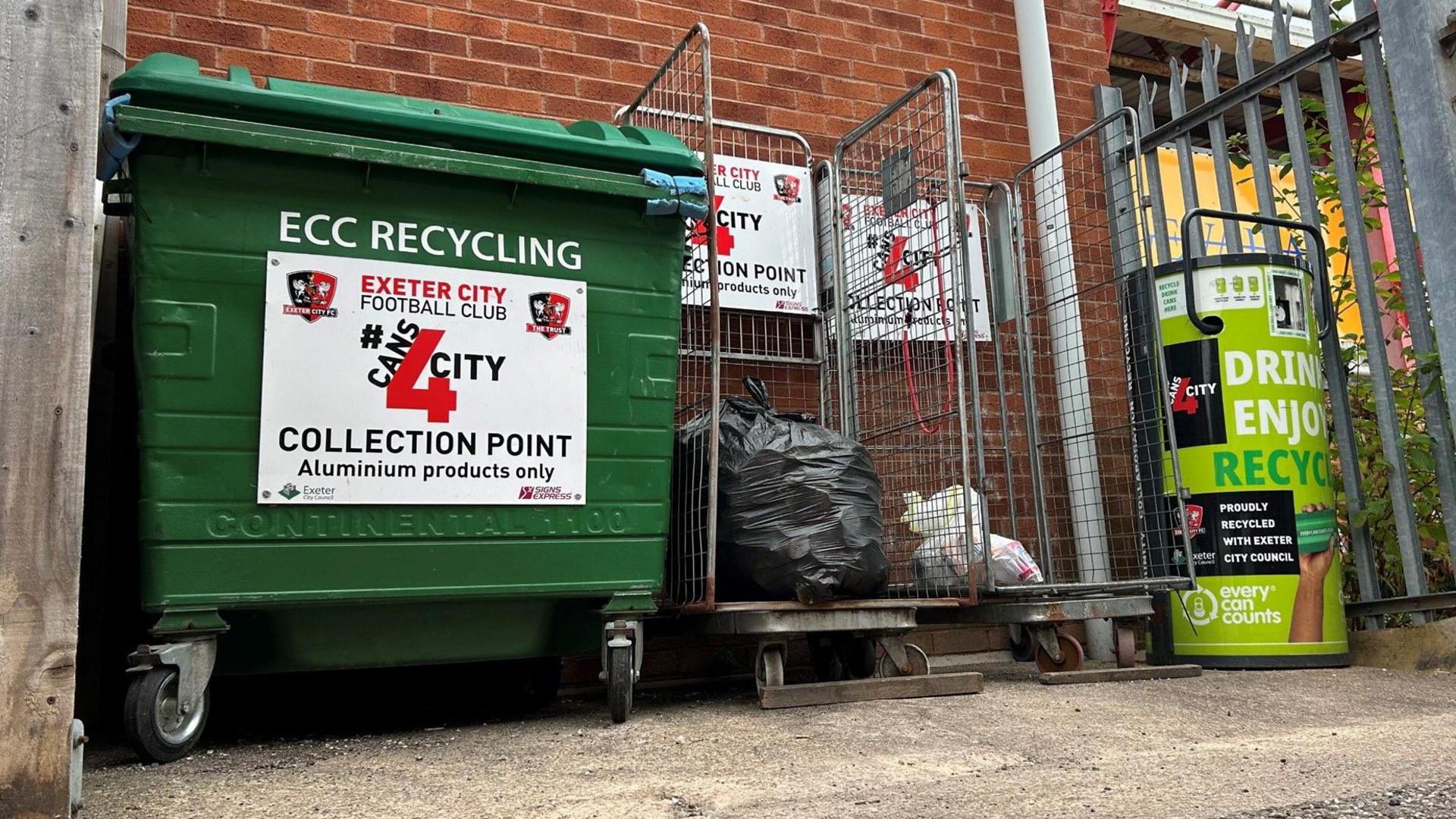 There is a large green bin as well as two metal cages with a black bin bag in one of them filled with empty crushed cans. There is also a can crusher with recycling branding next to the man. It's all located outside Exeter City Football Club.