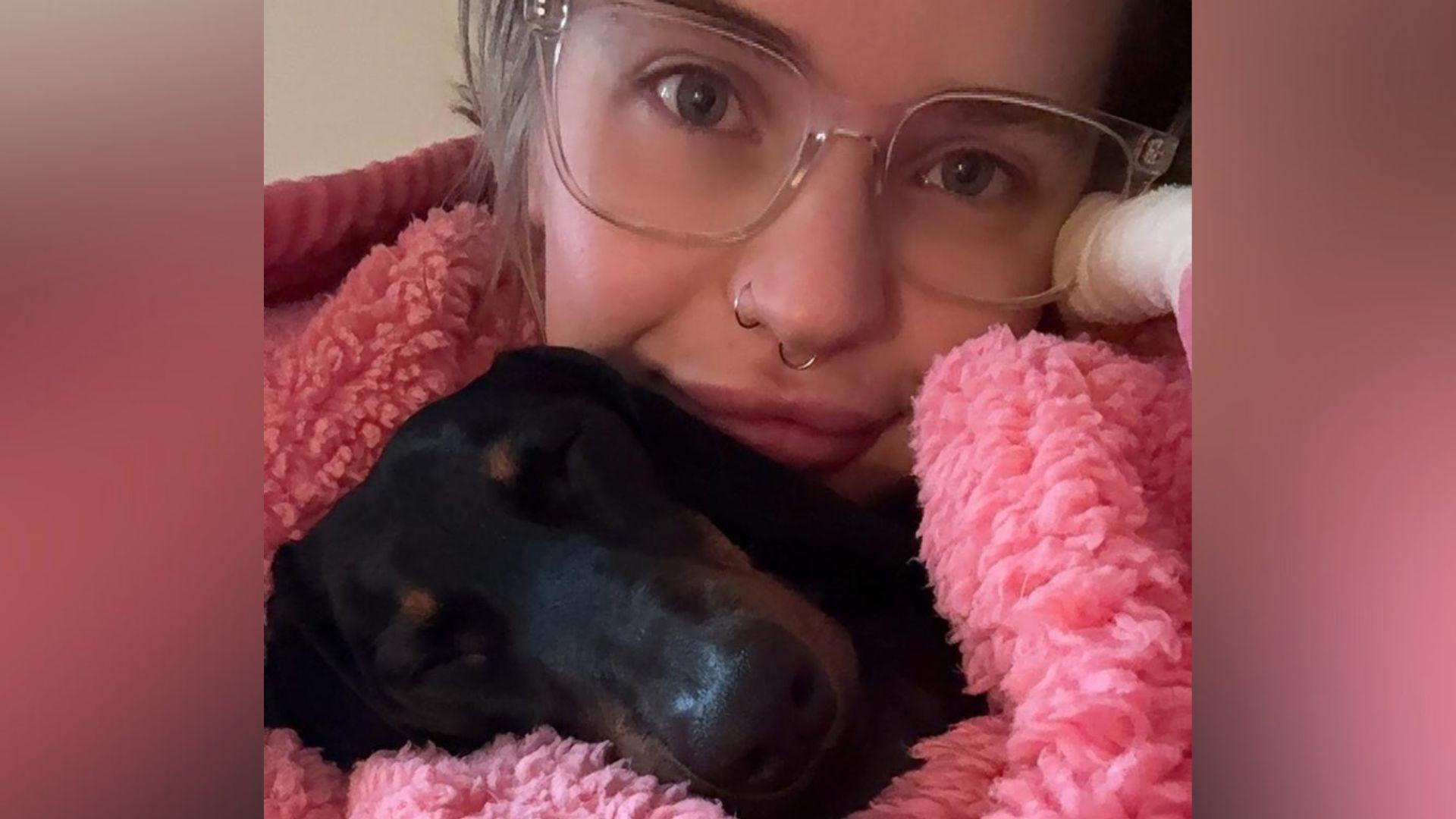 A close-up selfie of a woman with blond hair in a bun and two nose rings. She has a blanket or large jumper wrapped around her and a small black sausage dog is asleep on her chest.