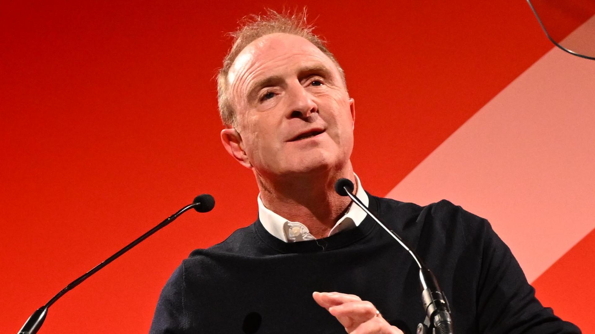 James Harding speaking at the Edinburgh TV Festival. He has short red hair and wears a white shirt under a thin black jumper. He has a neutral expression as he speaks into two small black microphones in front of a plain, bright red background.