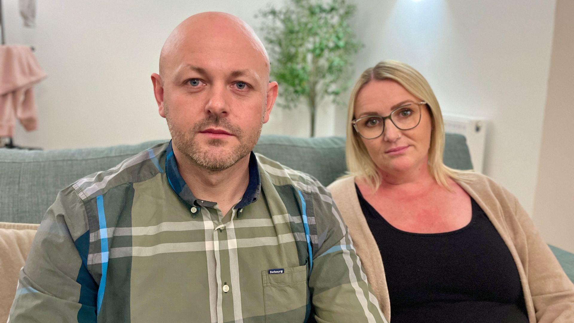 Chris McMaster and Natalie Davis sat on a turquoise sofa looking into the camera. Chris has stubble and is wearing a green checked shirt. Natalie has shoulder-length blonde hair and is wearing glasses and a beige cardigan over a black top.