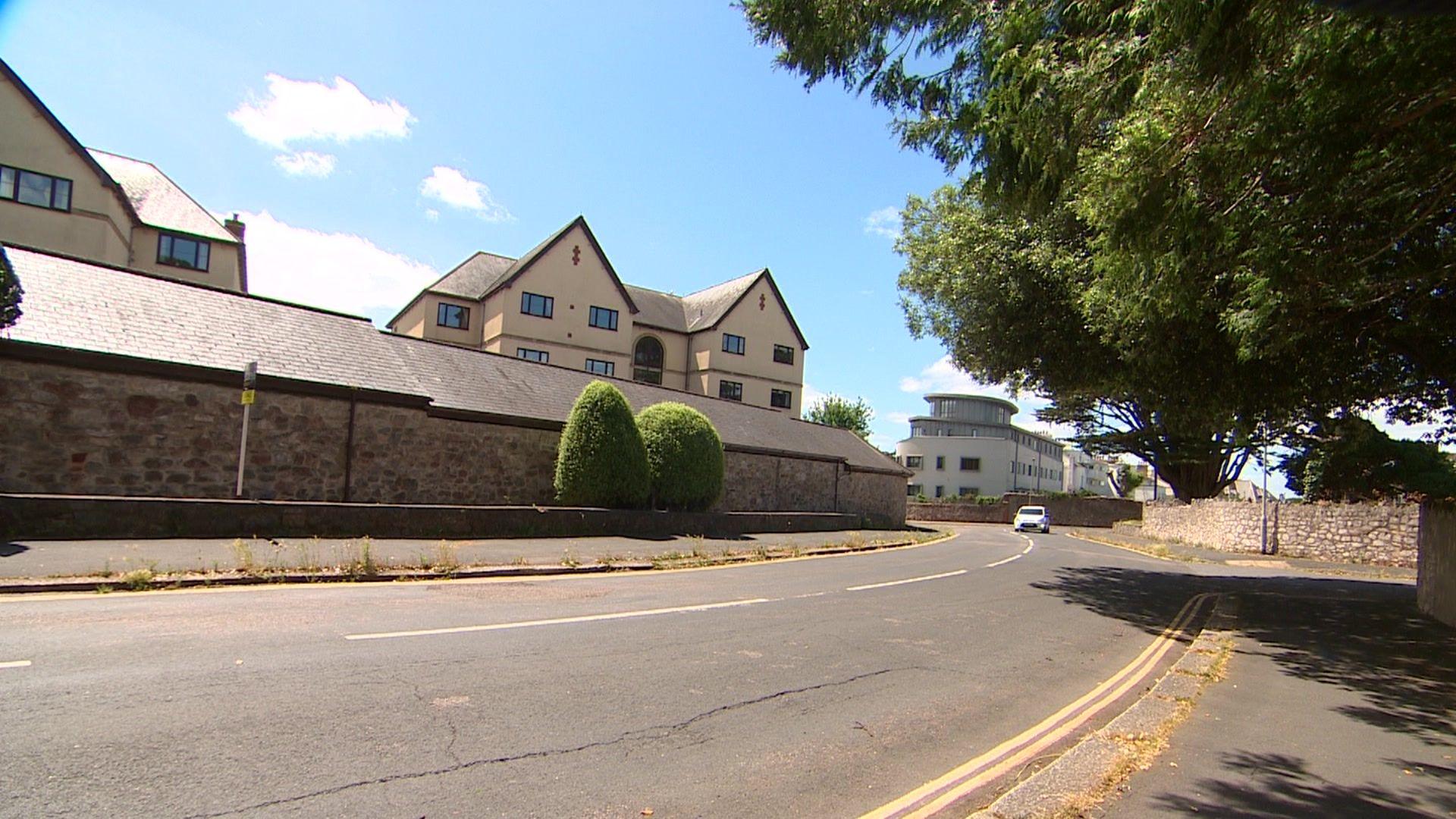 Blue skies over two-lane Douglas Road in the foreground with the buildings of the Deaf Academy in the background. A white car is in the distance travelling towards the camera