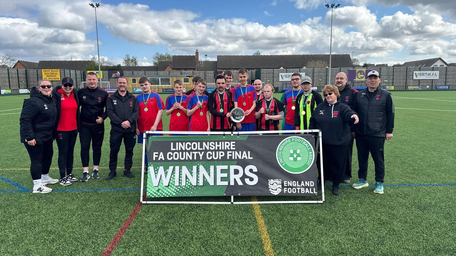 A line-up of players and coaching staff standing behind a placard which reads "Lincolnshire FA County Cup Final Winners".