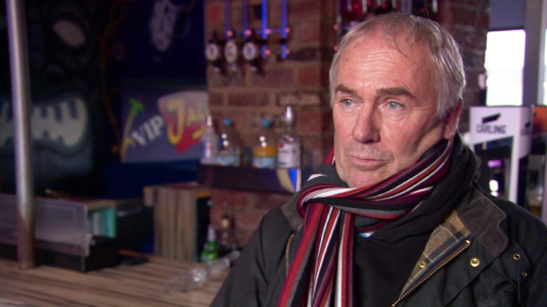 Mike Thompson, a man with short grey hair, is wearing a black jacket and a red, white and black scarf. He is standing in a bar with a wooden counter, bottles and beer taps.