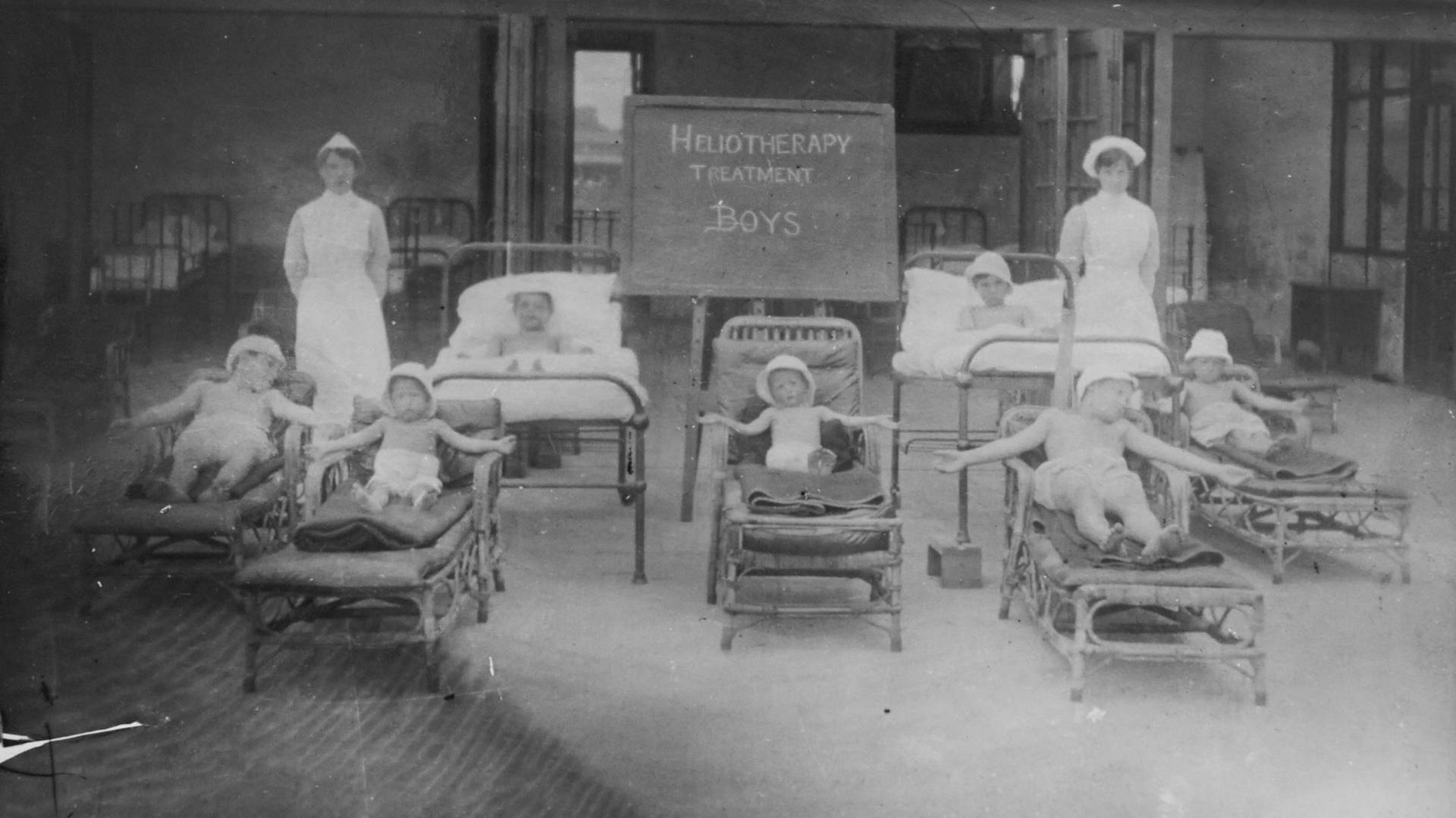 A black and white photo of several children on beds wearing shorts and caps with two nurses behind them.