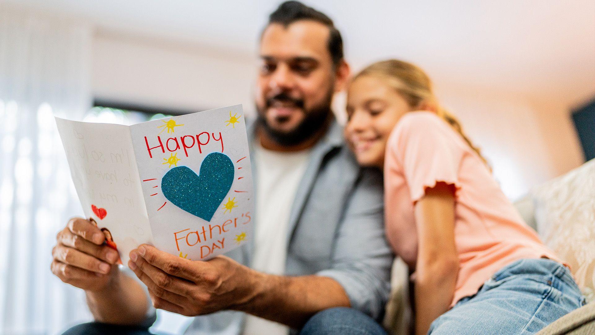 A blurred Dad and daughter are cuddled together. The Dad is holding a card which is in focus in the foreground. It says Happy Father Day with a big glittery heart in the middle