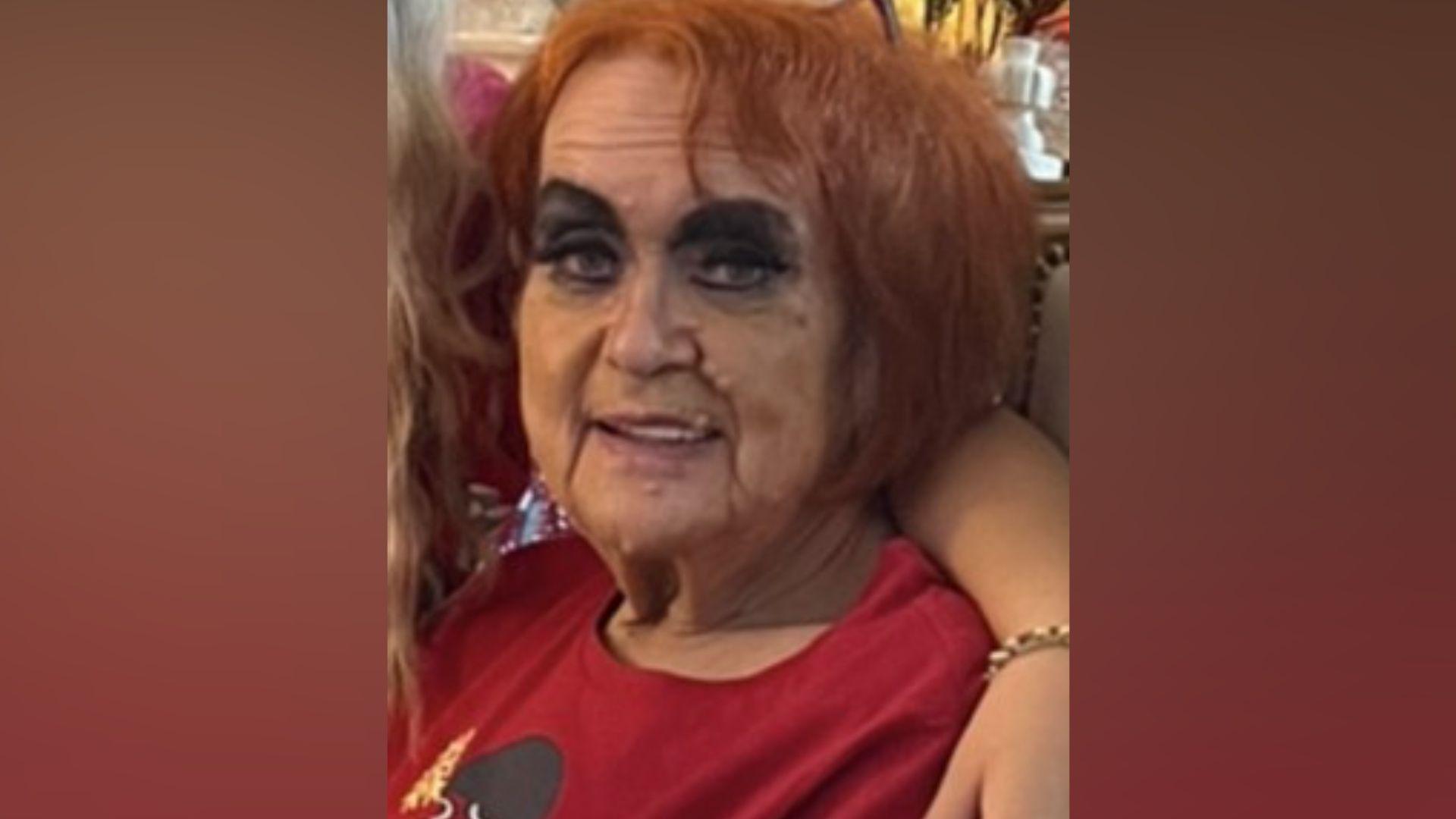 A woman with auburn hair and black eyeliner on is wearing a red top. Someone has an arm around her.