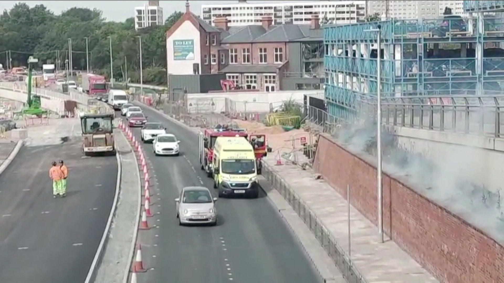 Cars and lorries drive along a dual carriageway. A fire engine and a yellow emergency service vehicle are parked in the righthand lane. A cloud of smoke can be seen on a grassed area. A blue multi-storey car park is visible in the background.