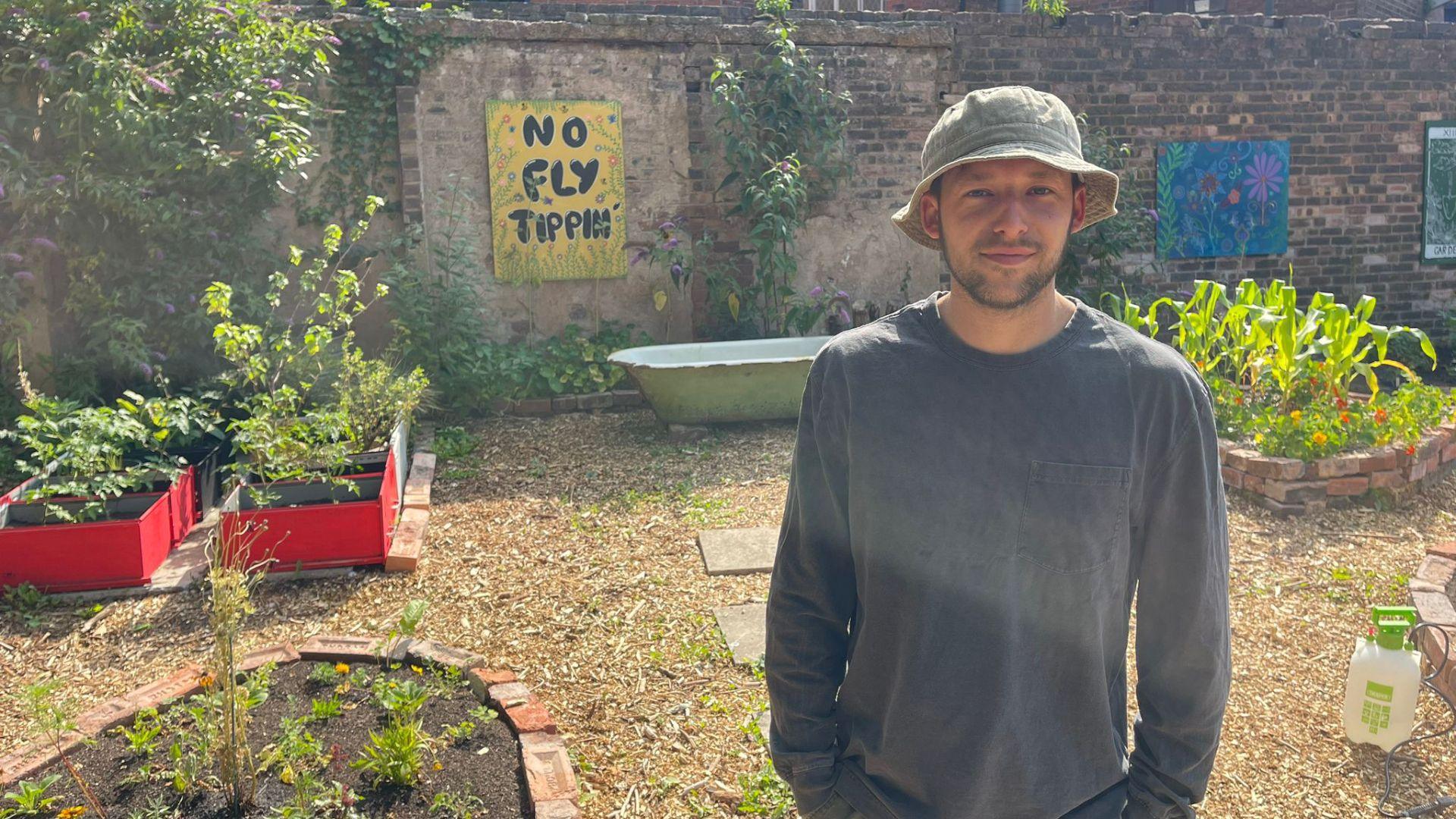 A man in an olive bucket hat and grey jumper stands with his hands in his pockets in a garden. The garden has beds of plants made with bricks and old filing cabinets. There are also paving slabs amongst the wood chipping garden surface, a bath and a mechanical watering can. There are brick walls bordering the garden with flowery artwork and a sign that says 'NO FLY TIPPIN'