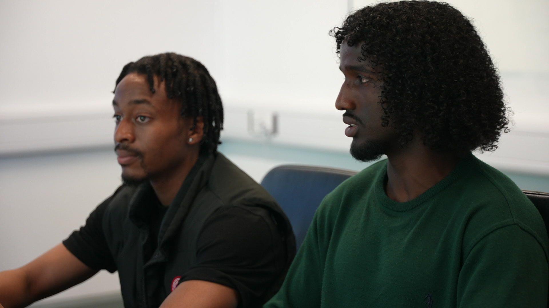 Tosin, 20, and Adnan, 18, sit at the table in an office room taking part in the discussion. Tosin, wearing a black sleeveless jacket, looks straight ahead, listening, while Adnan, in a dark green top, is speaking.