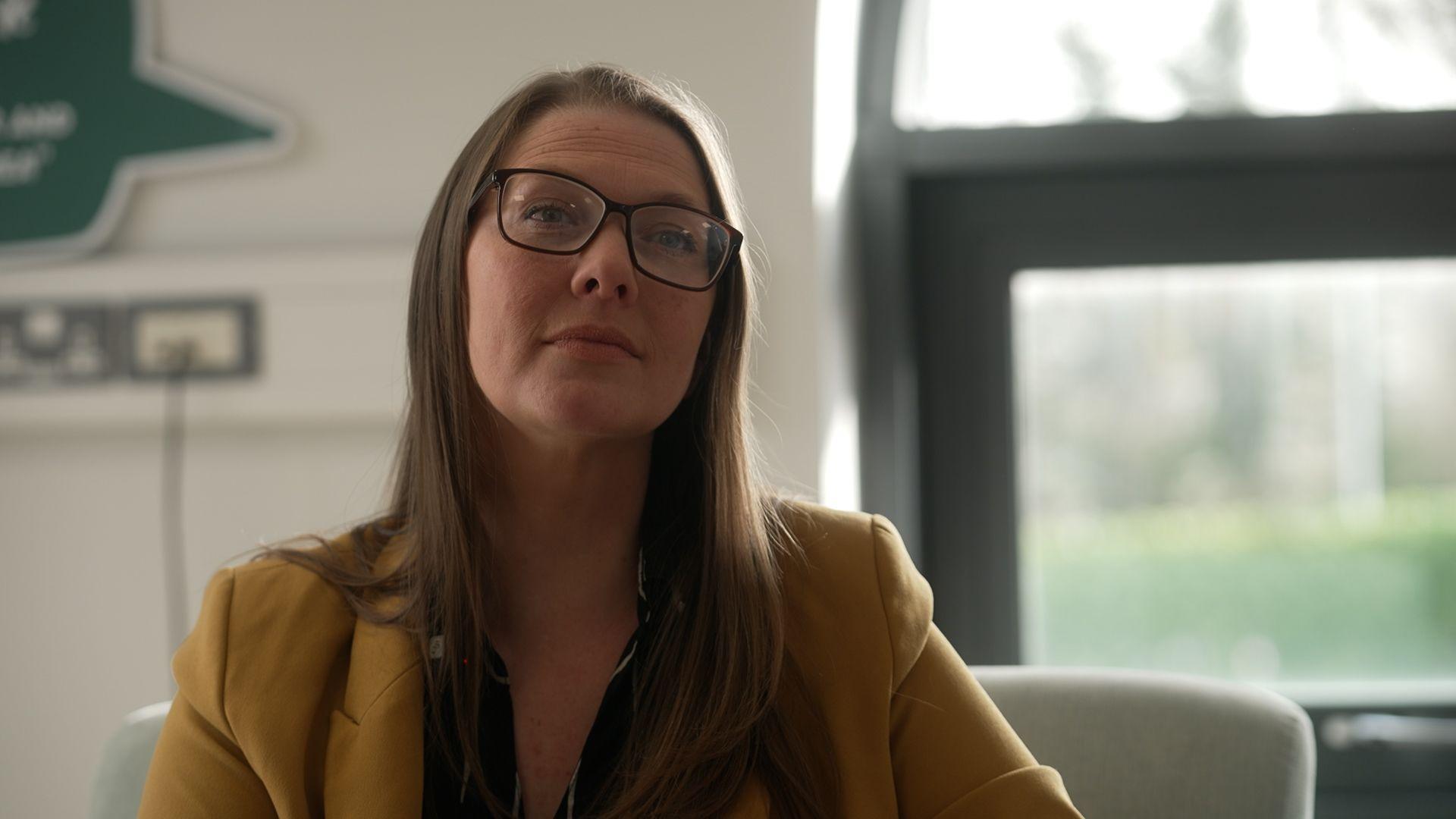 A woman wearing glasses and a yellow blazer looks into camera, seated on an armchair in a white office with a bright window behind her.