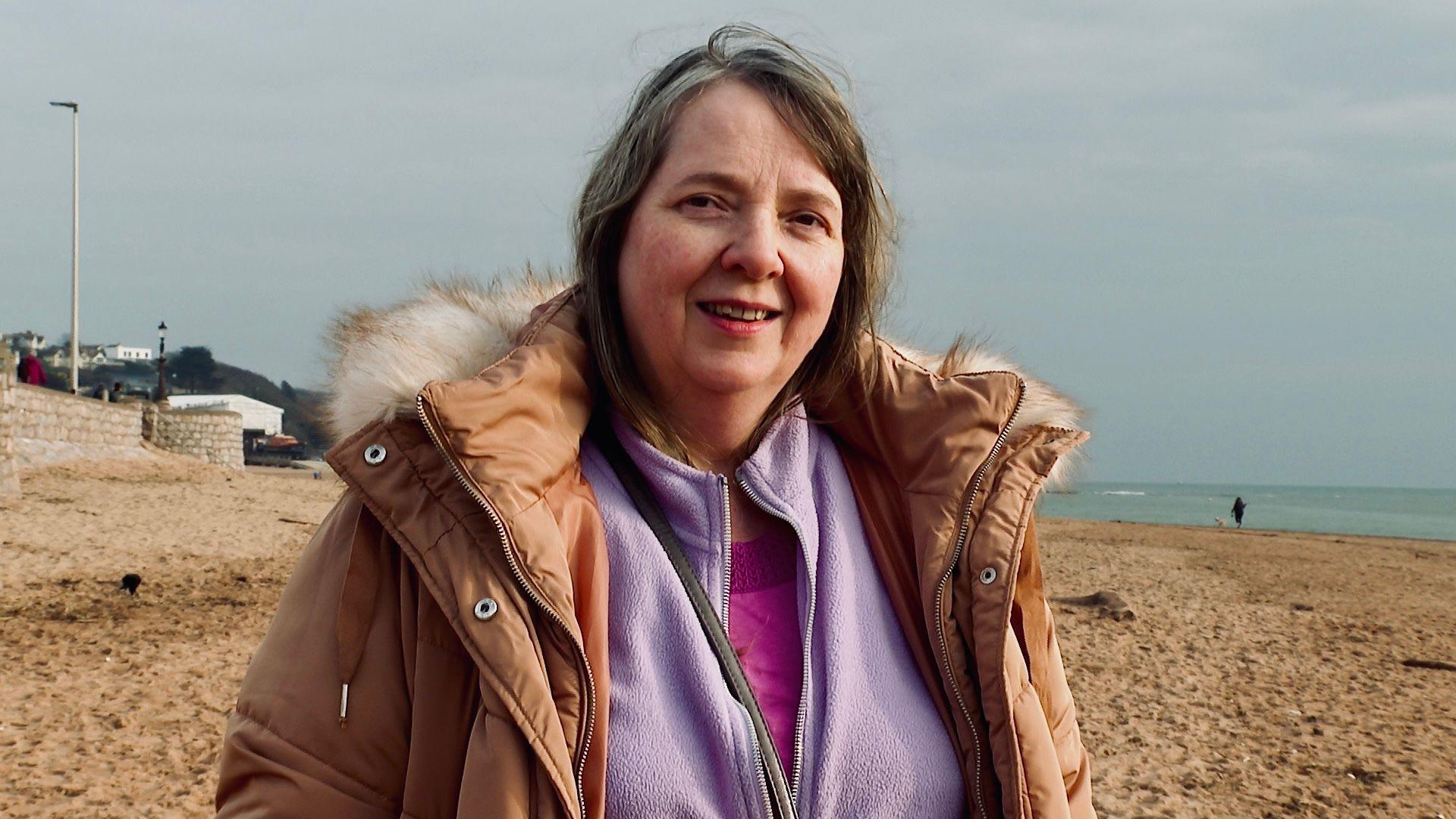 Claire Lambert, an unpaid carer, wearing a pink top, a lilac fleece and a beige coat with a fluffy collar, on the beach at Exmouth