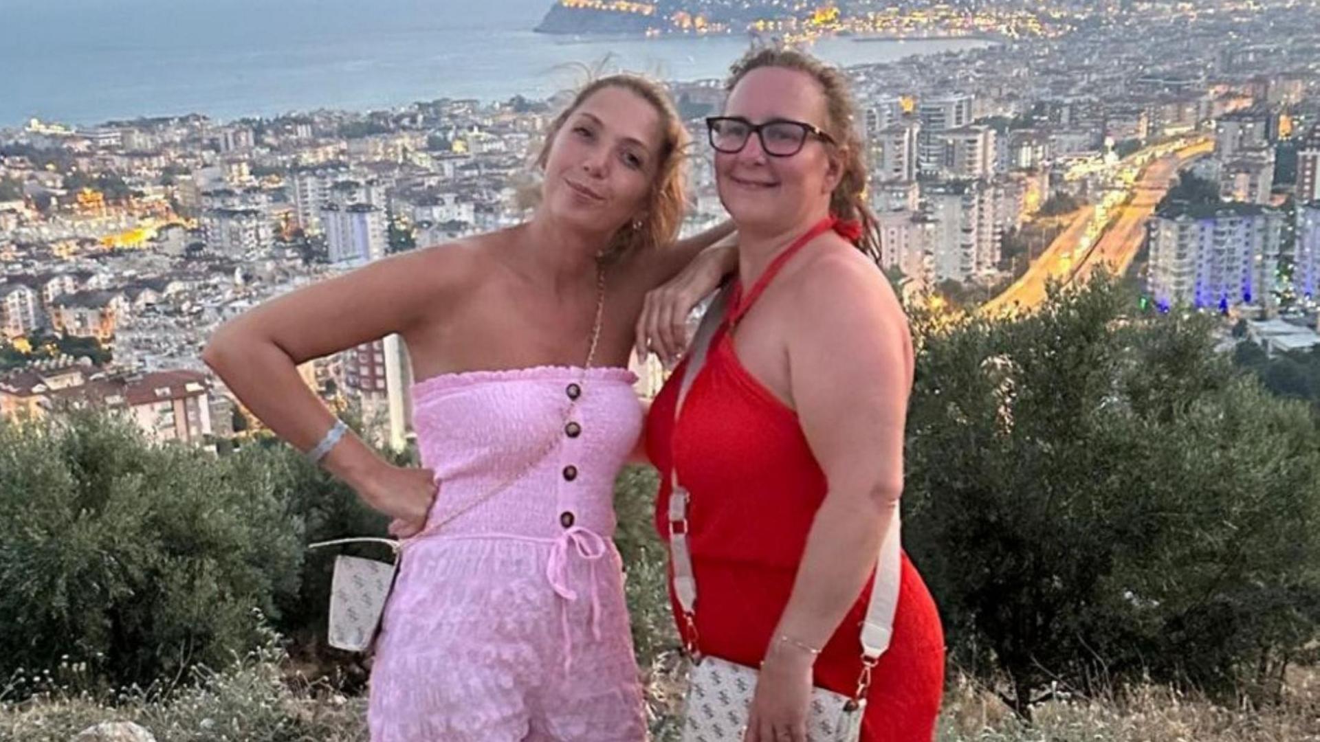 Two woman. On wearing pink, the other red. They are smiling stood on a hill. Behind them is Alanya in Turkey.