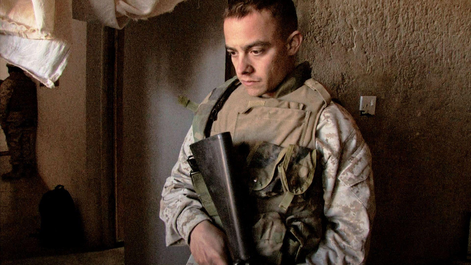 A man in camouflage clothing with a khaki vest over the top holds a gun and looks down at the ground. Behind him is a rough beige wall. 