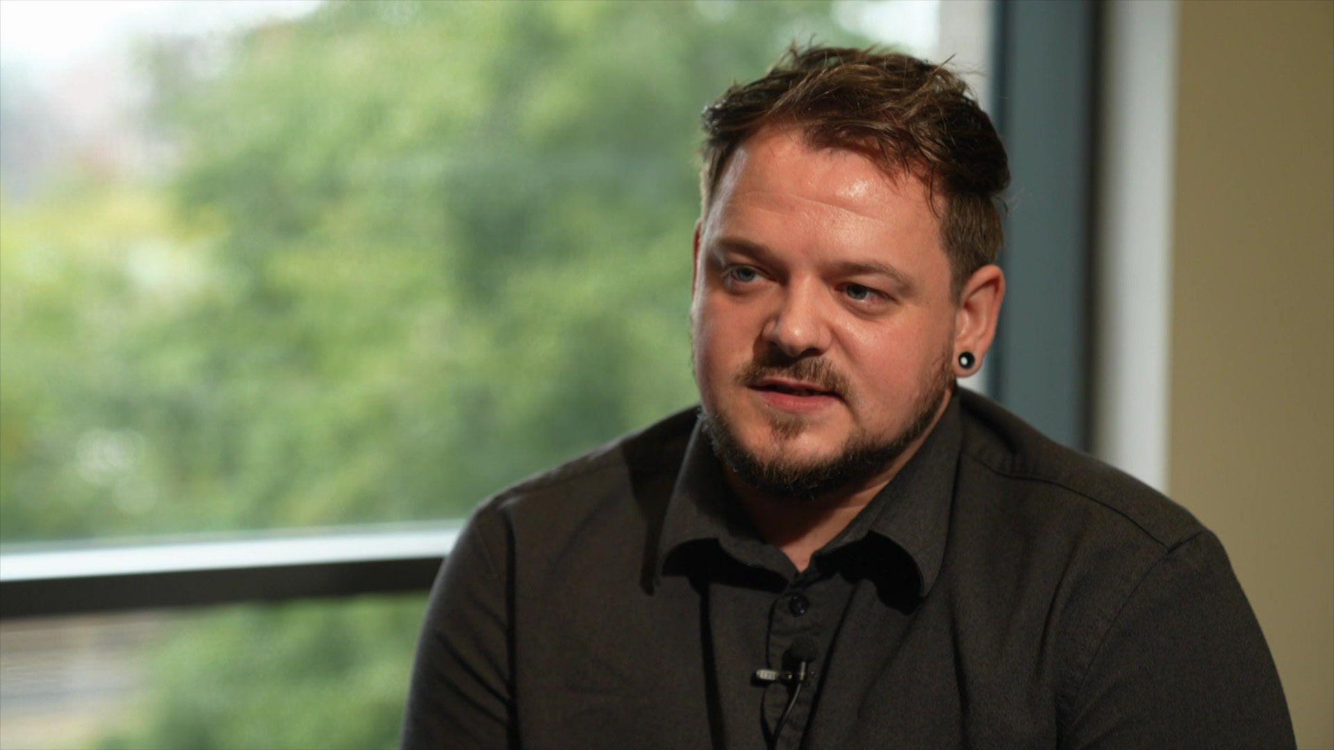 Man in a black shirt with an earring sitting near a window above a green space.