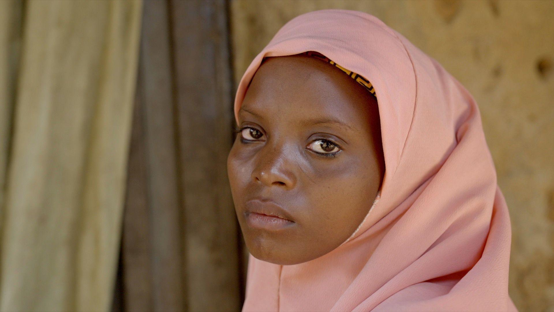 Aisha, with her hair covered, looks at the camera